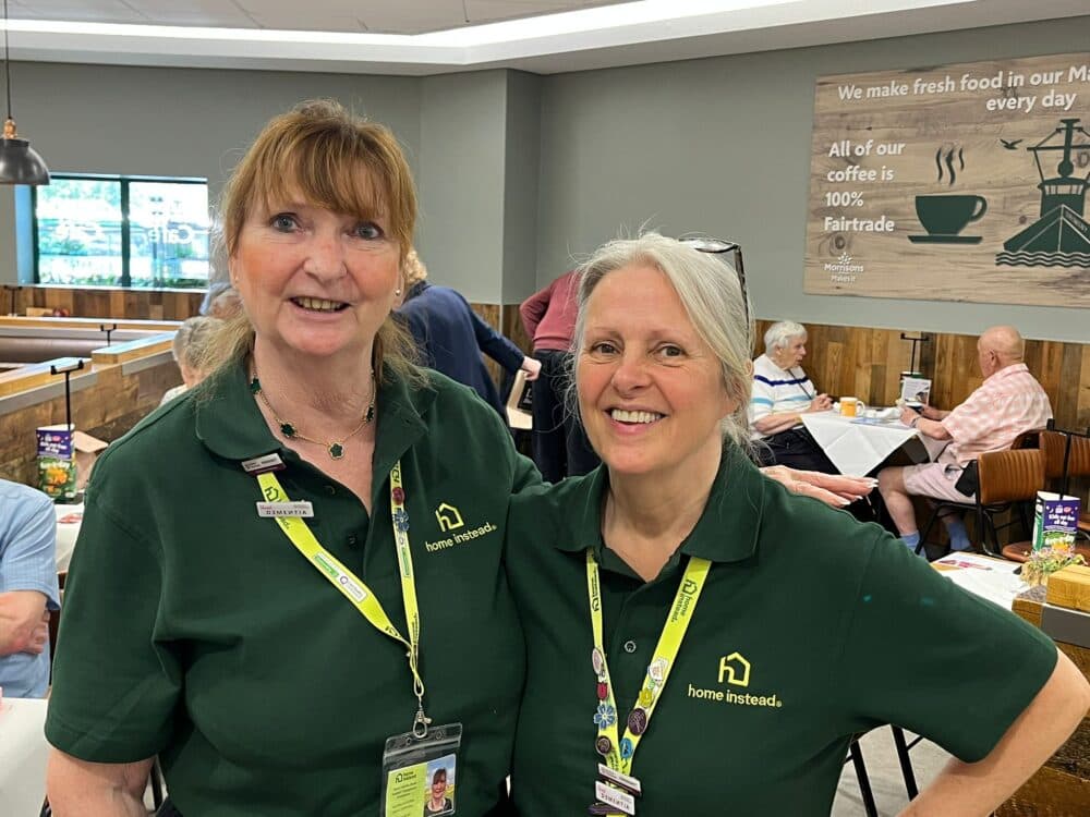 Two smiling women wearing green "home instead" shirts stand together in a bright, busy café. - Home Instead