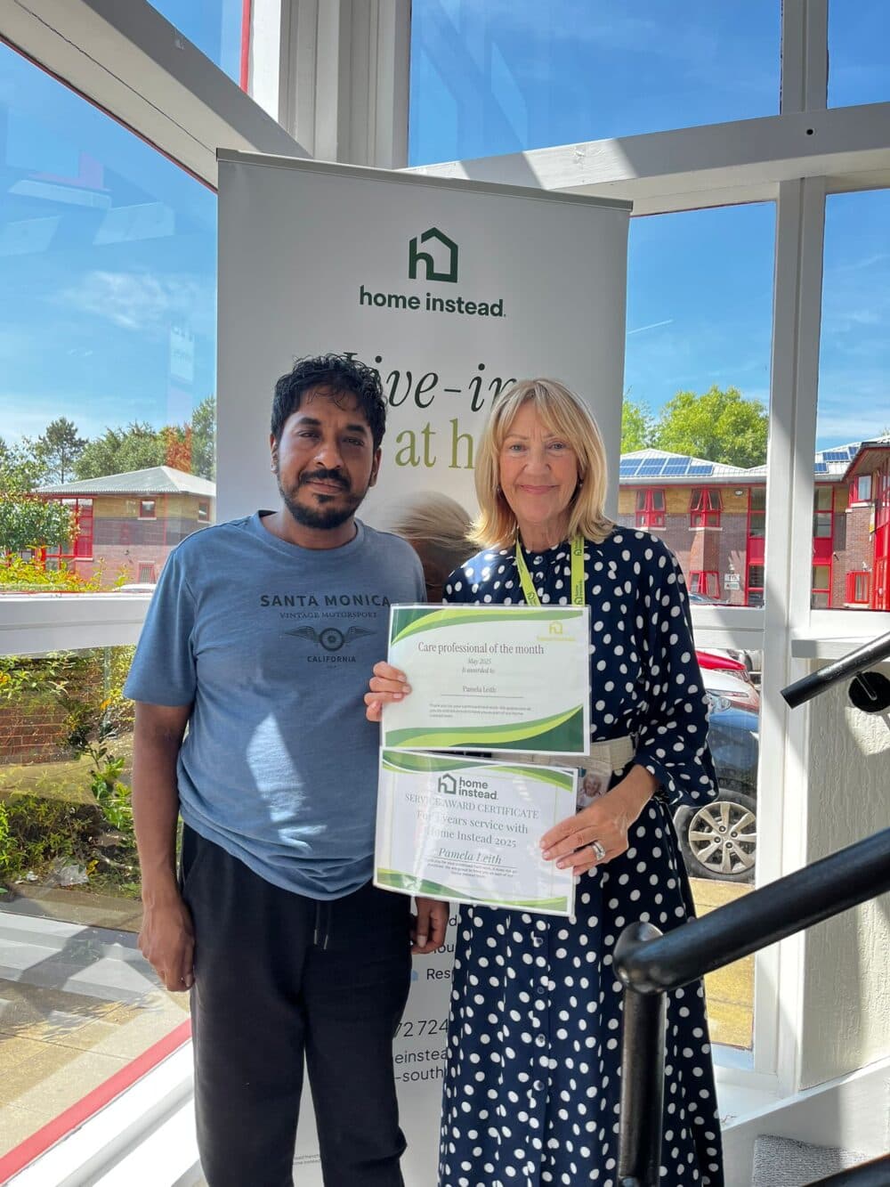 Two people stand indoors; one holds certificates in front of a "Home Instead" banner and large windows. - Home Instead