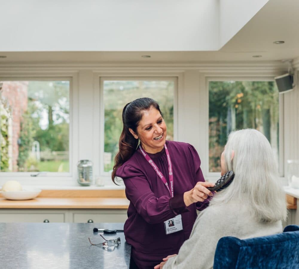 A carer wearing maroon and with long black hair combing the hair of an older adult inside a home with lots of windows