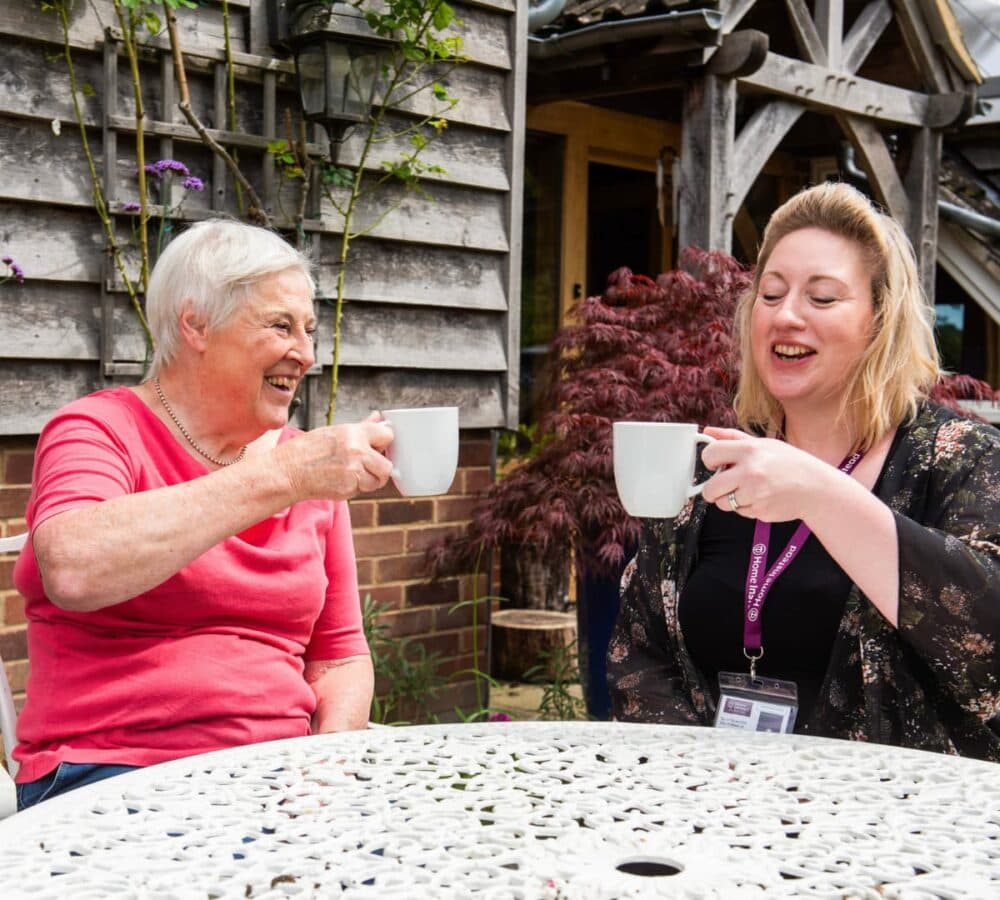 Two women sit outside at a table, smiling and holding mugs, enjoying a drink together. - Home Instead