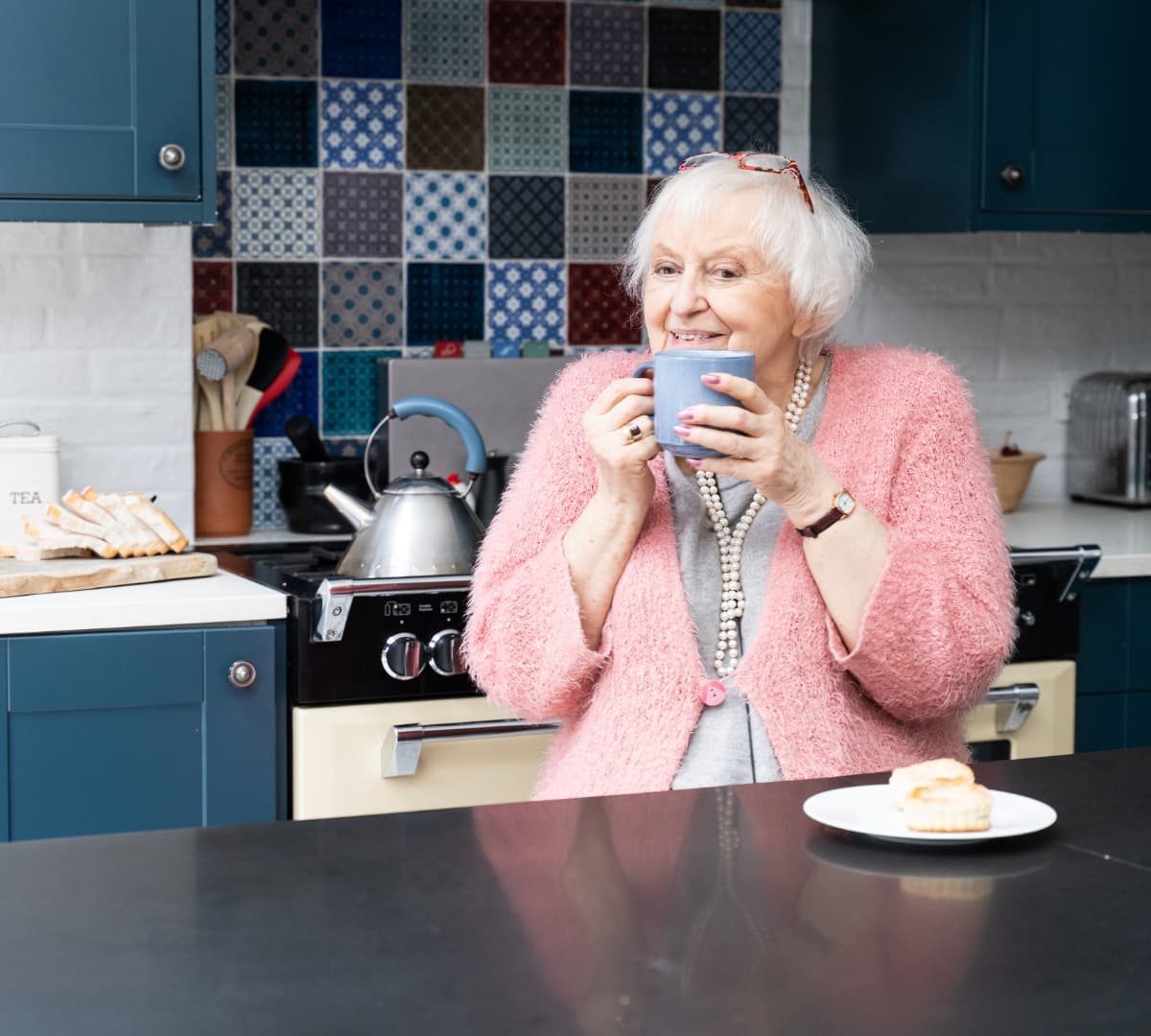 Happy senior woman with short white hair smiling and sipping a cup of tea in he kitchen while wearing a pink sweater