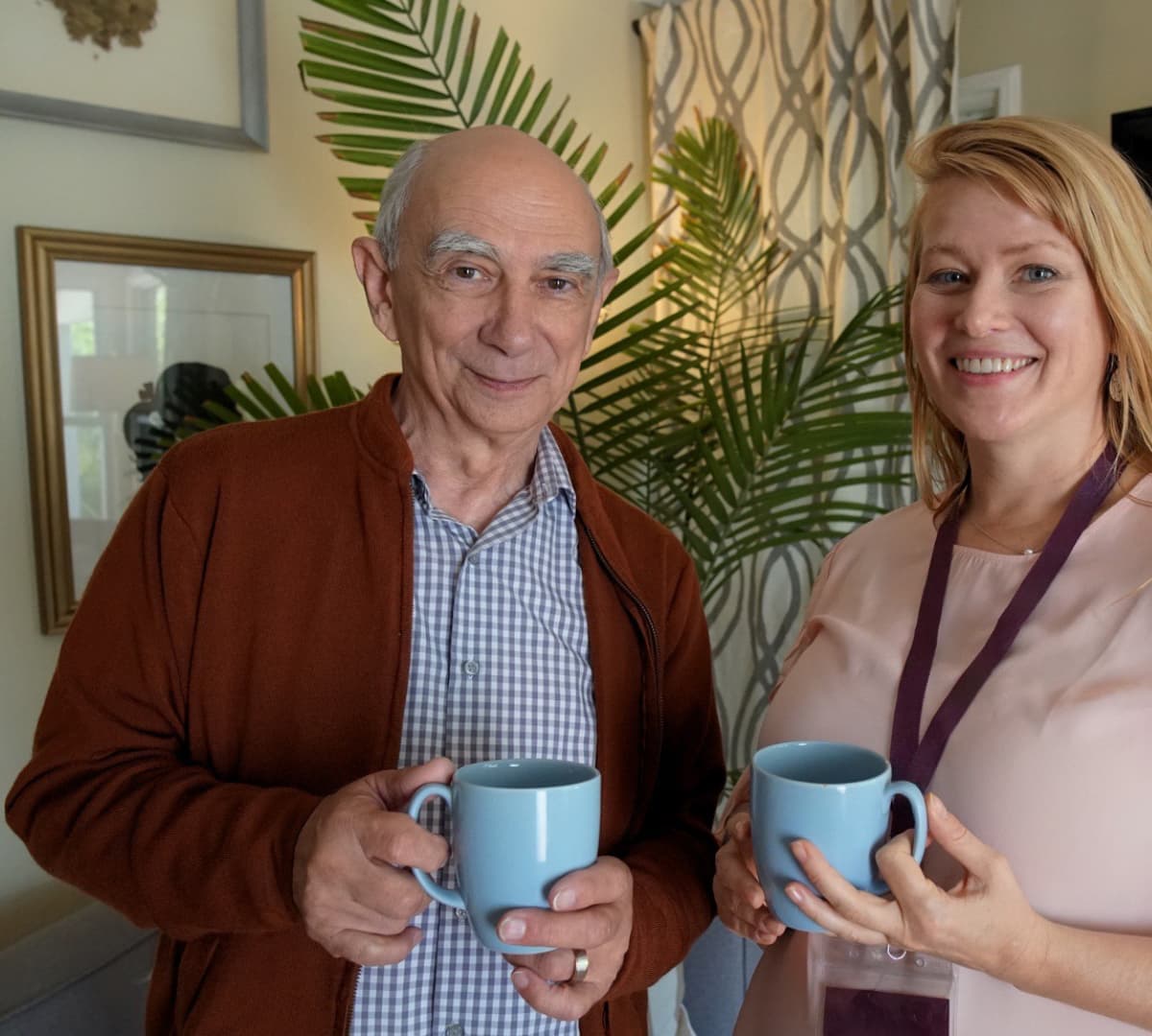 Old man with white hair standing with his carer with long hair while holding a cup of coffee in blue mugs