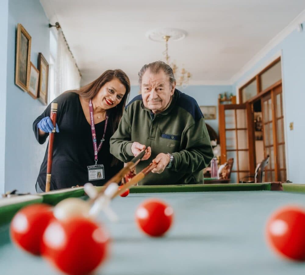 Older man with grey hair playing billiard inside a room with his carer with long hair playing with him