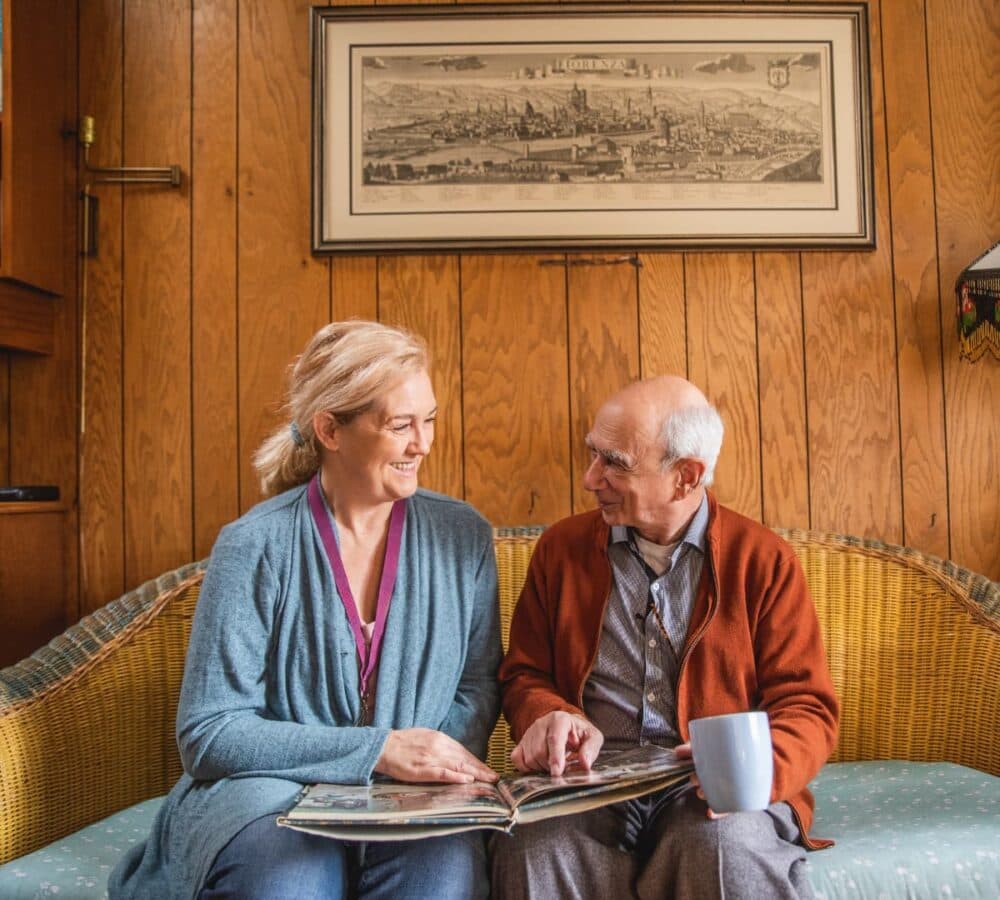 Older man with white hair sitting on a couch inside his home while holding a cup and looking at a photo album with his lady carer wearing blue and both smiling