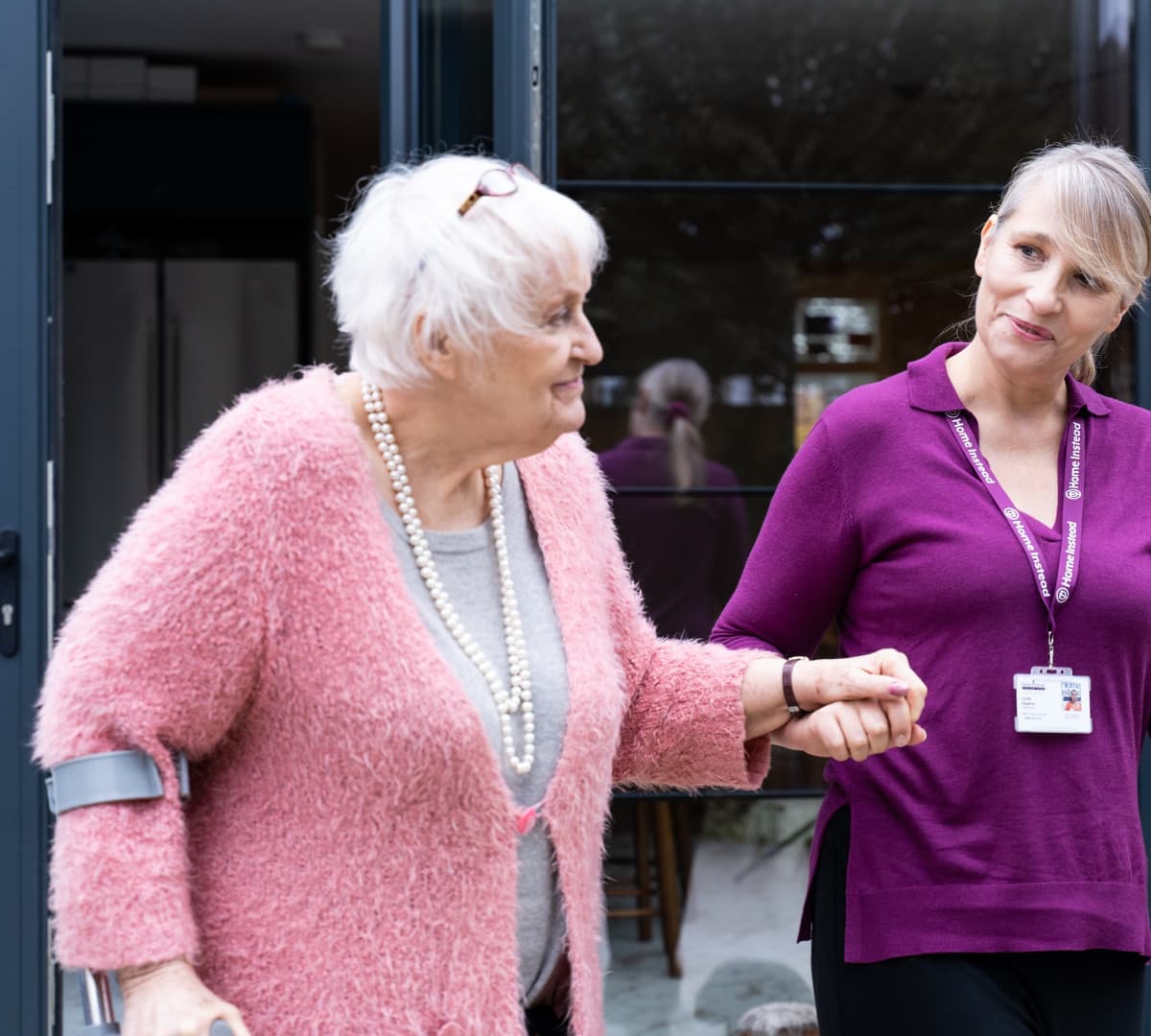 Older woman wearing pink sweater with white wait walking while being assisted by her carer