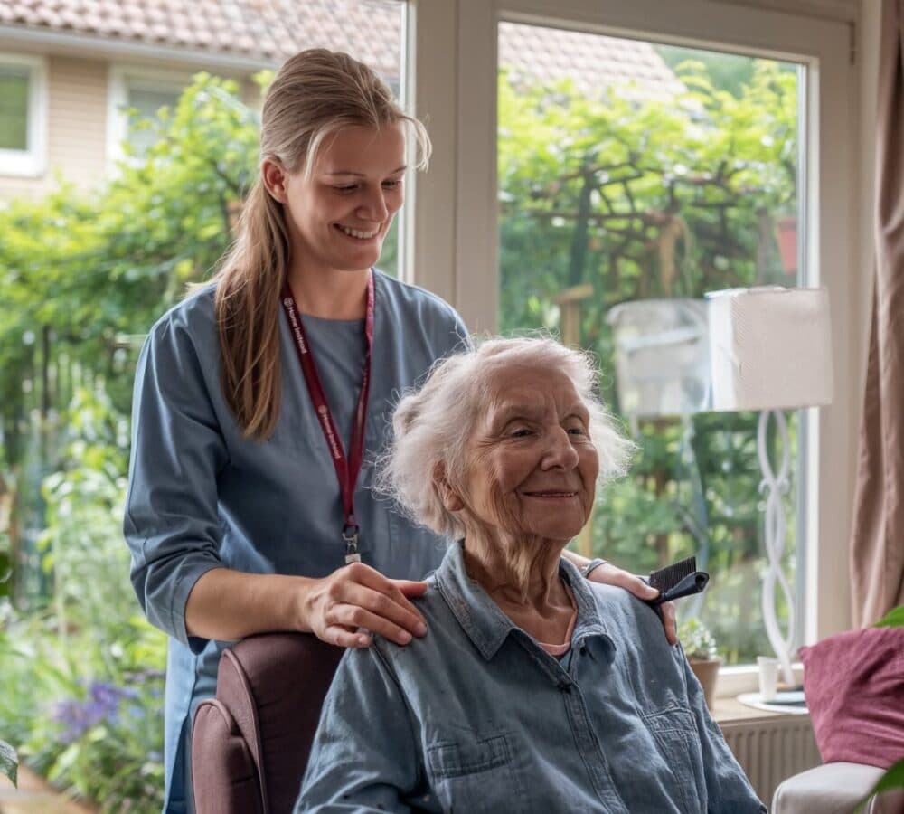 Senior woman sitting on a chair with short white hair while her carer is standing at her back with long hair smiling while combing the hair of the senior woman with glass window at the back