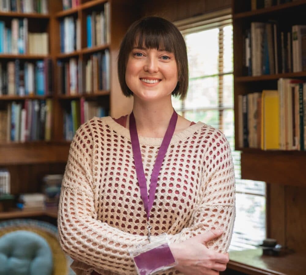 Woman with short brown hair smiling, wearing a white knit sweater and lanyard, standing in a library. - Home Instead