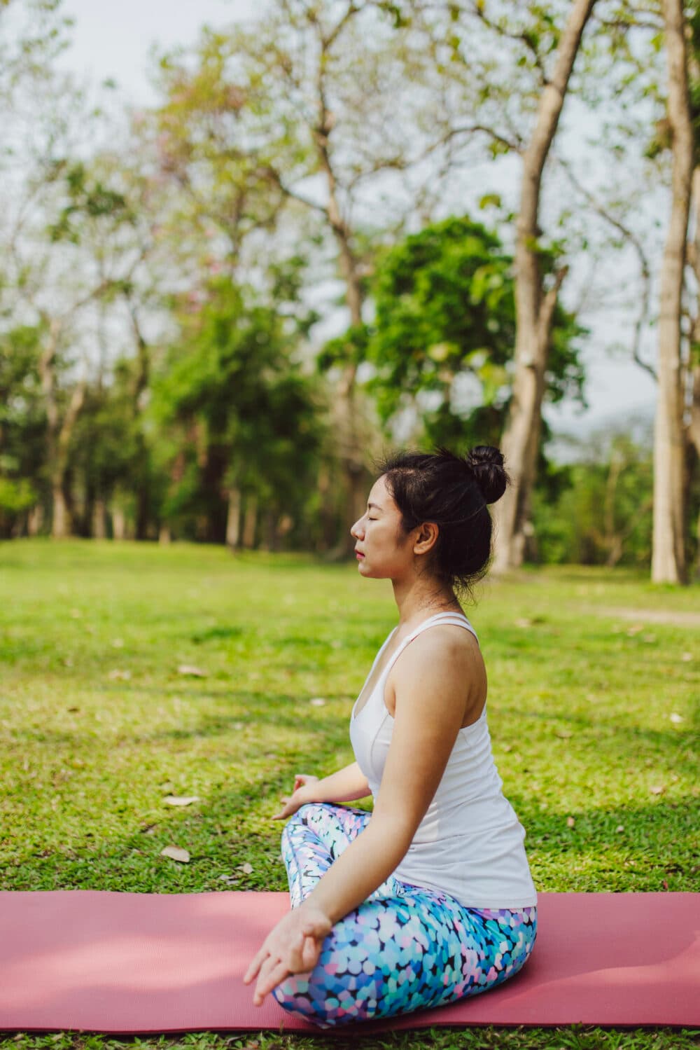 Woman meditating on a yoga mat outdoors, sitting cross-legged in a green park with trees in the background. - Home Instead