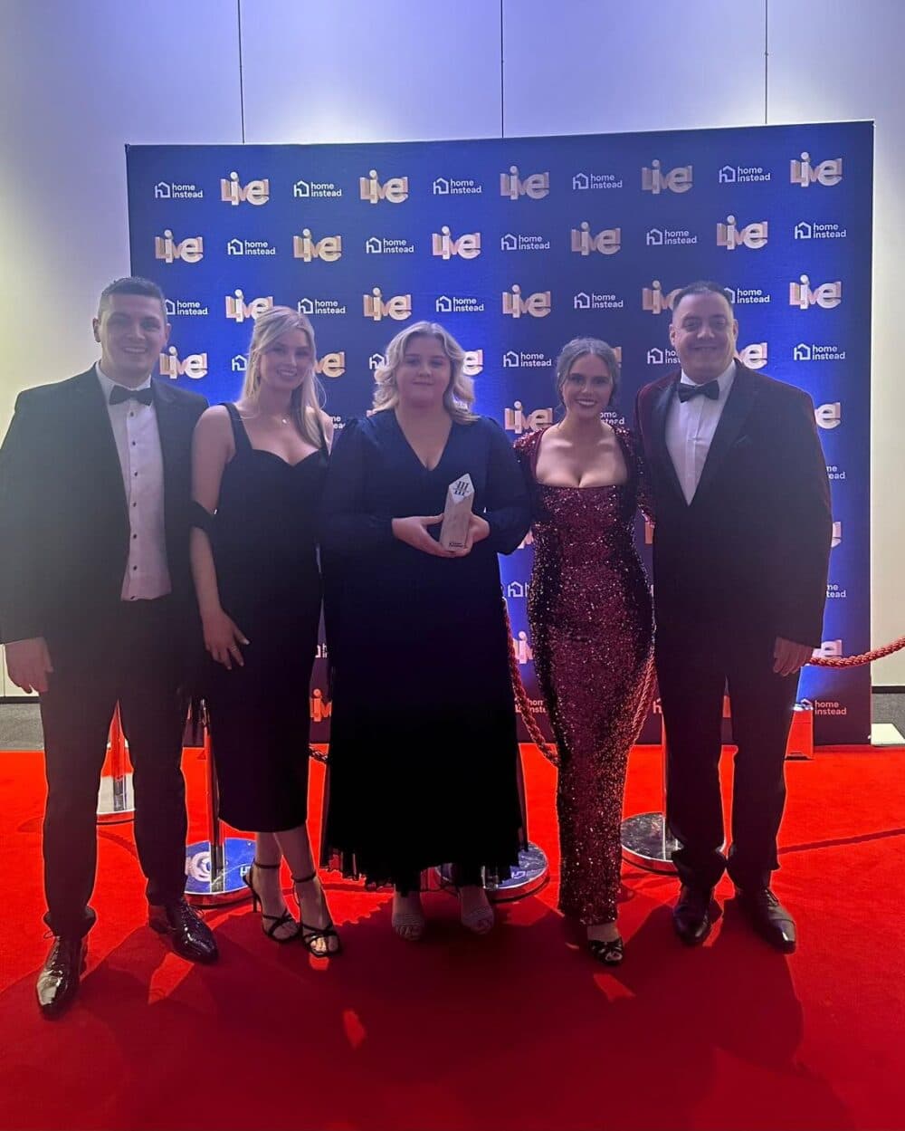 Five people dressed formally stand on a red carpet, posing with an award in front of an event backdrop. - Home Instead