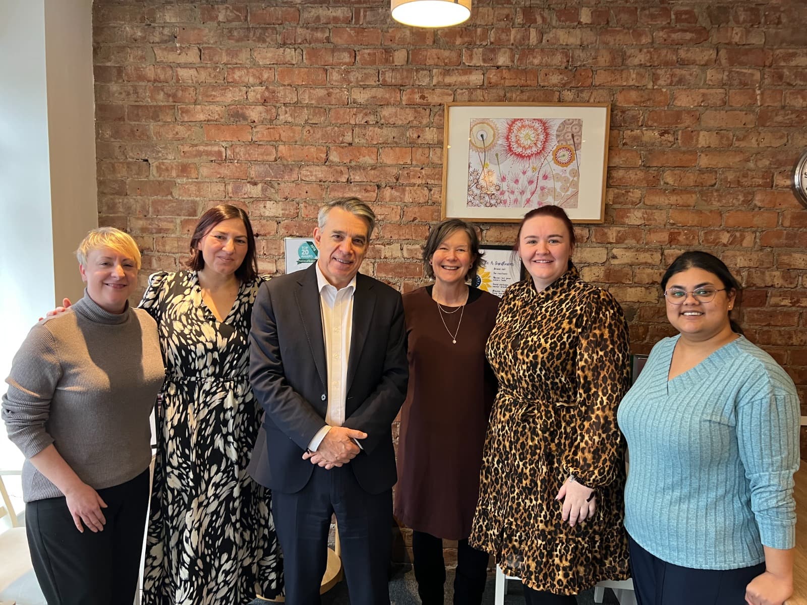 Six people stand smiling together indoors in front of a brick wall with artwork hanging behind them. - Home Instead