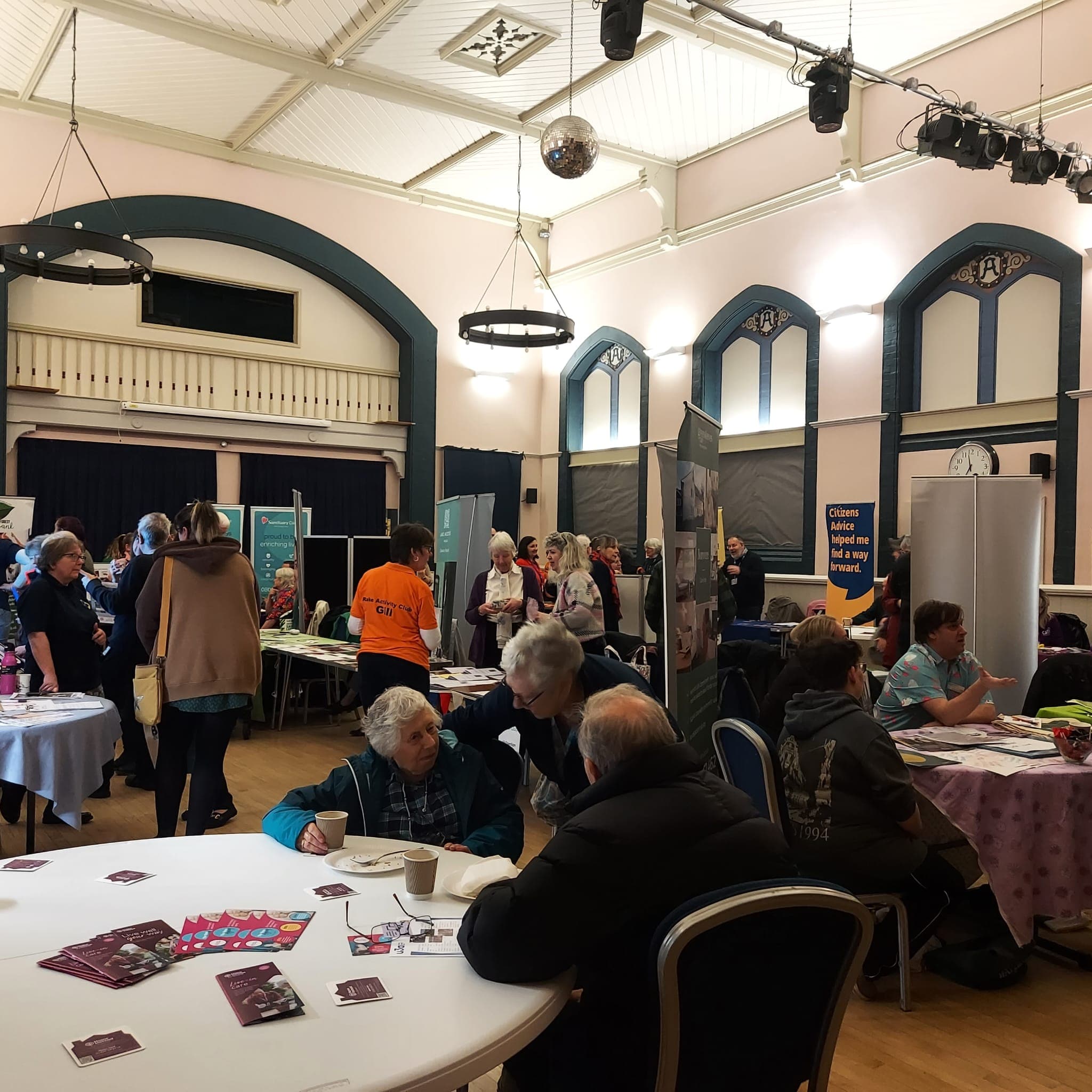 People attending an indoor community fair, talking at tables and visiting informational booths in a hall. - Home Instead
