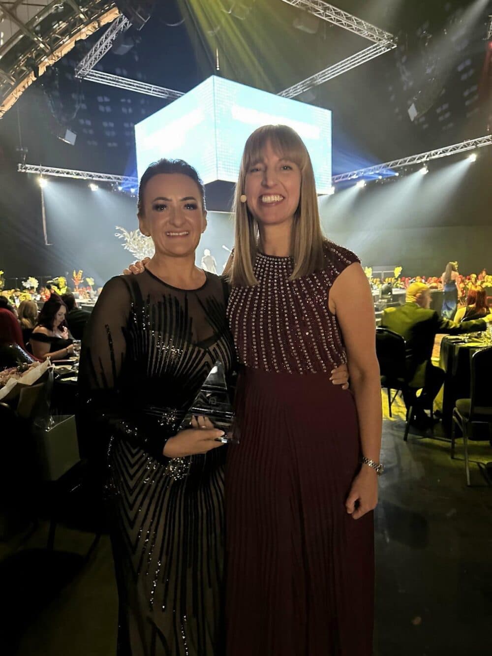 Two women in formal dresses smile at an event with tables and a large screen in the background. - Home Instead