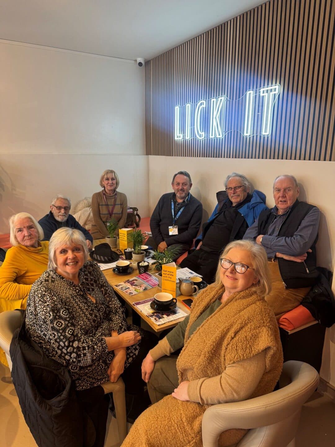 Eight older adults sit around a café table with drinks under a neon “LICK IT” sign on the wall. - Home Instead