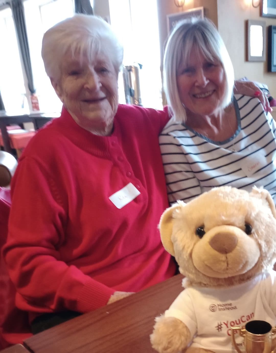 Two older women smiling at a table with a teddy bear mascot in front of them in a cozy room. - Home Instead