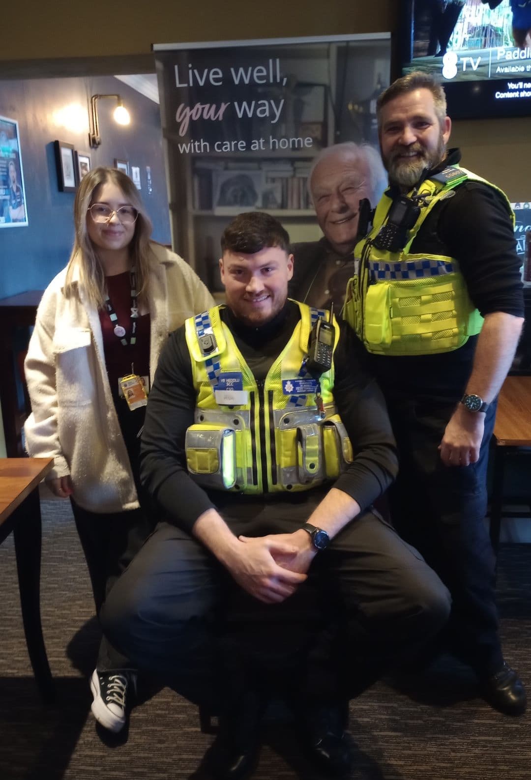 Three people, including two police officers, pose and smile in front of a care-at-home promotional banner. - Home Instead
