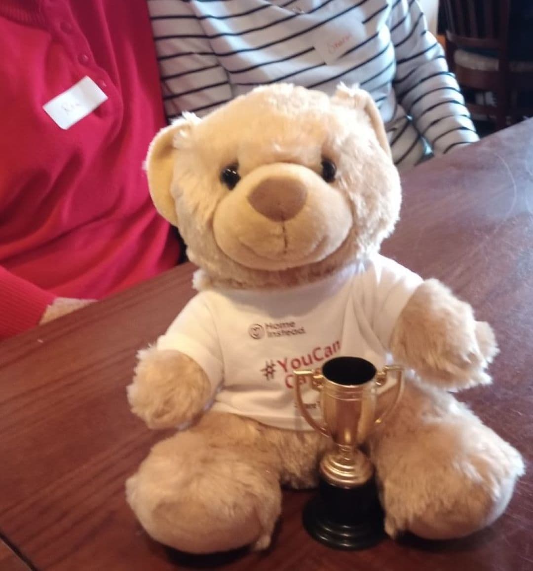 A teddy bear in a white shirt holding a small trophy, seated at a wooden table with people in the background. - Home Instead
