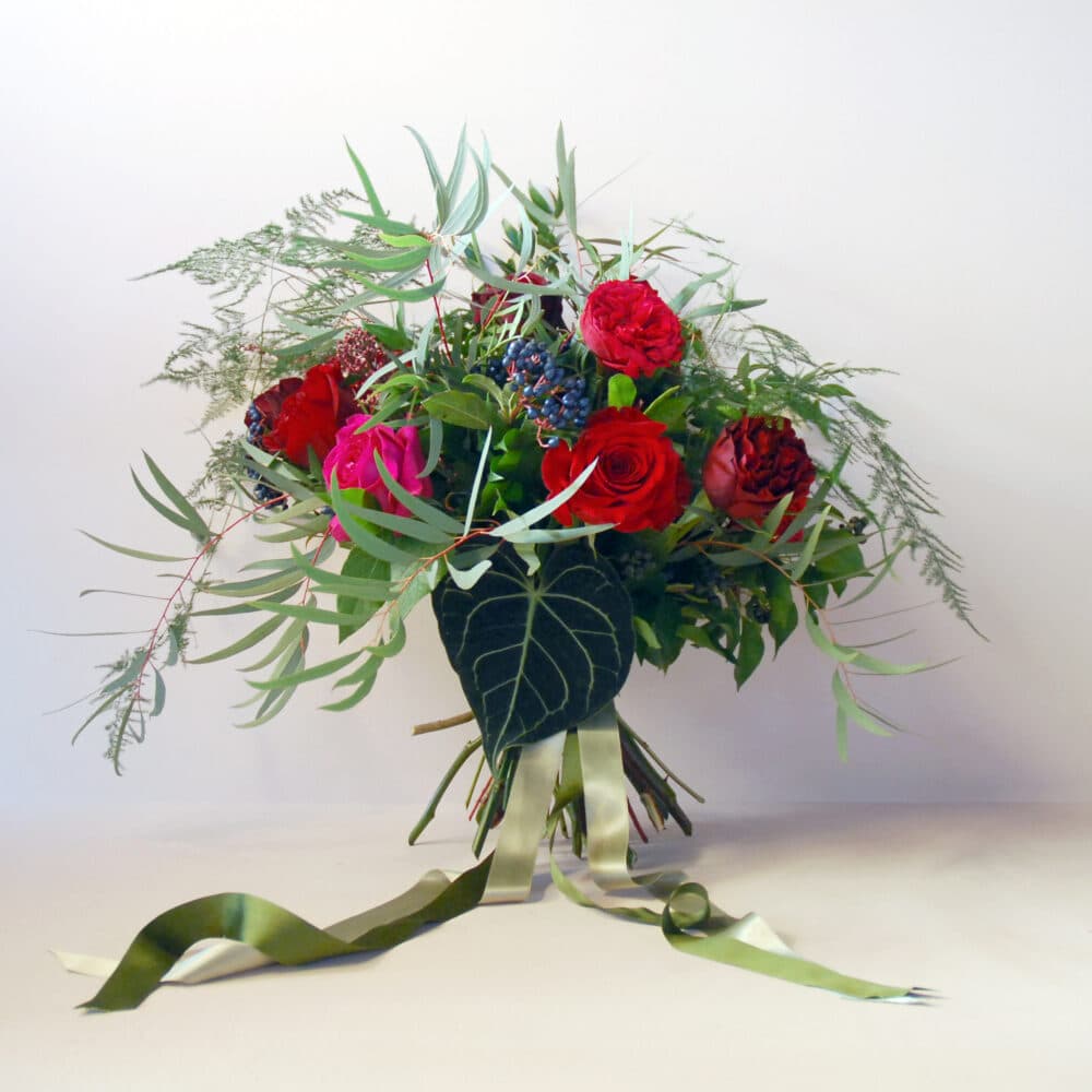 A beautifully arranged bouquet featuring deep red and bright pink roses, clusters of dark berries, and lush greenery, including eucalyptus and delicate ferns. The bouquet is accented with a striking dark green anthurium leaf at the base and tied with flowing green and ivory ribbons. Set against a neutral white background.