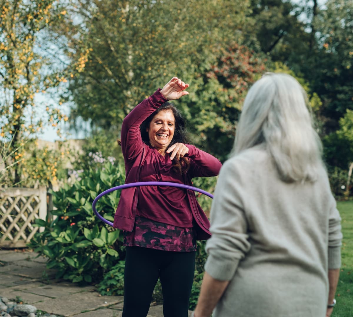 Care Professional wearing maroon outside the garden while playing hula hoop with a female older adult client