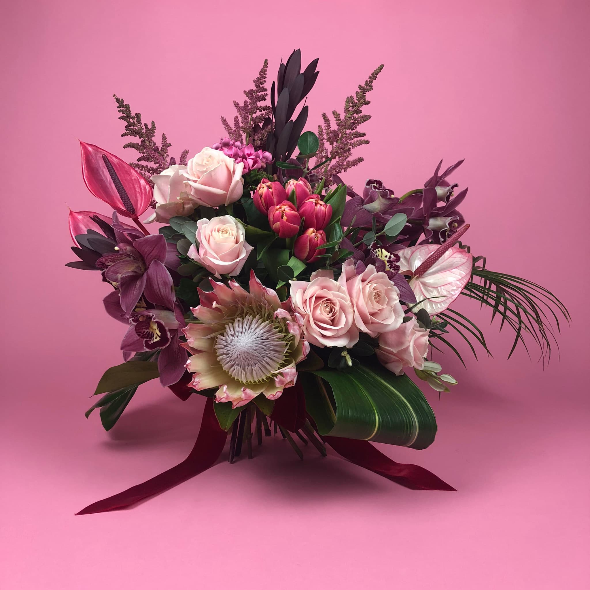 A luxurious bouquet of flowers arranged against a pink background. The bouquet features a mix of soft pink roses, red tulips, dark purple orchids, and a striking king protea at the center. Complemented by lush green foliage and exotic leaves, the arrangement is tied with a deep red ribbon.