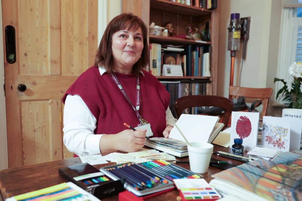 Woman sitting at a table with art supplies, sketchbook, and colored pencils, smiling at the camera. - Home Instead