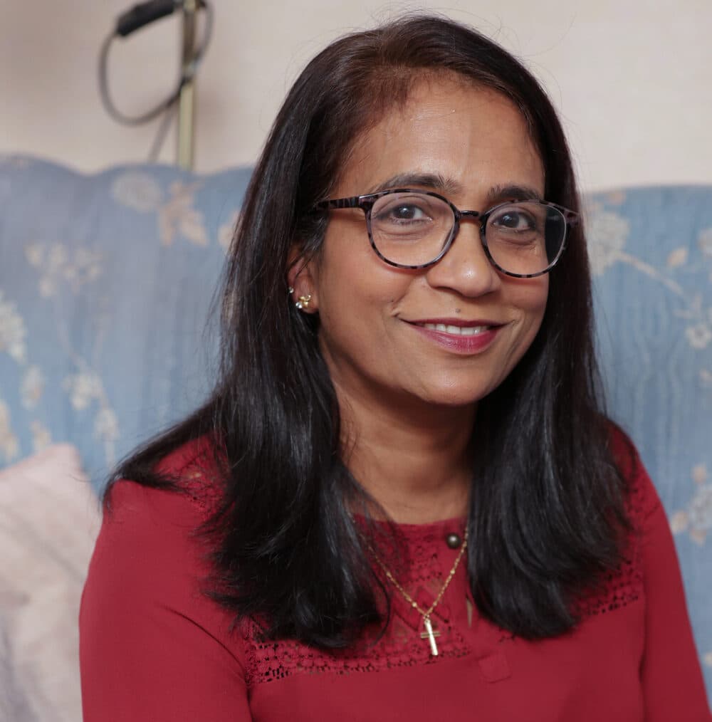 Woman with glasses and long dark hair, wearing a red blouse, smiling while seated on a blue patterned sofa. - Home Instead