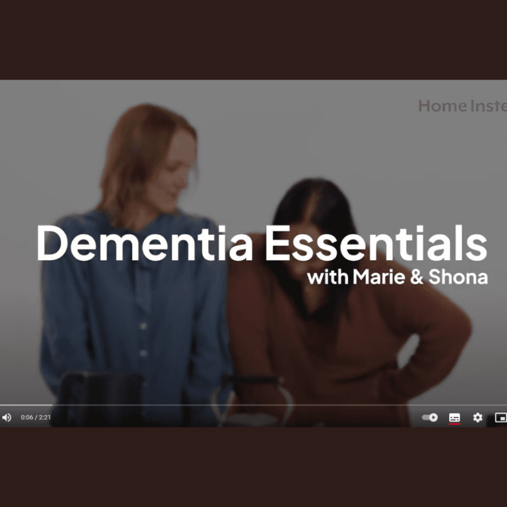 Two women stand side by side under the title "Dementia Essentials with Marie & Shona" on a video screen. - Home Instead