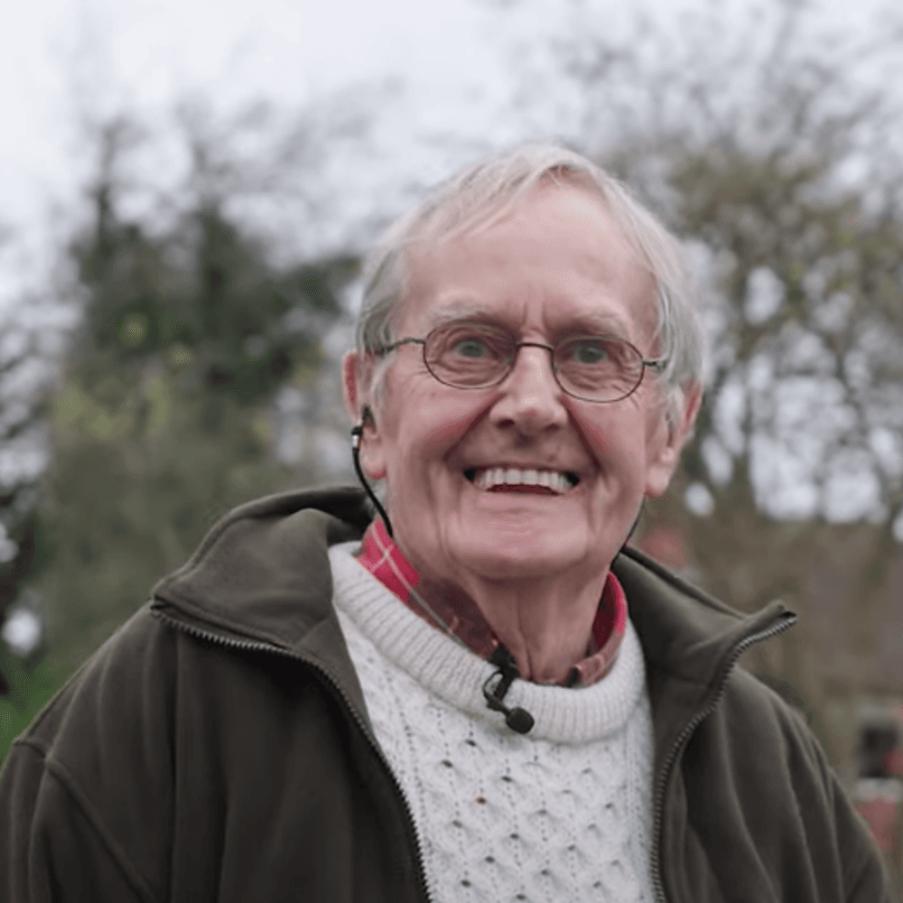 An elderly man with glasses smiles outdoors, wearing a sweater, plaid shirt, and jacket. - Home Instead