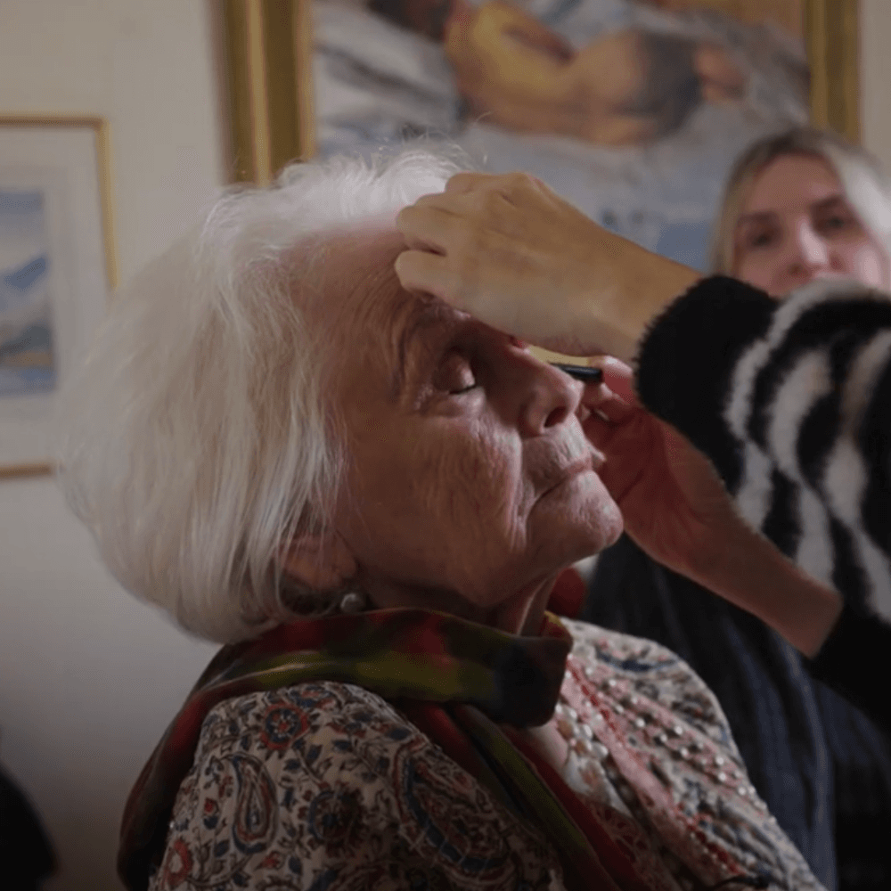 An elderly woman with closed eyes gets her makeup done by someone in a striped sweater. - Home Instead