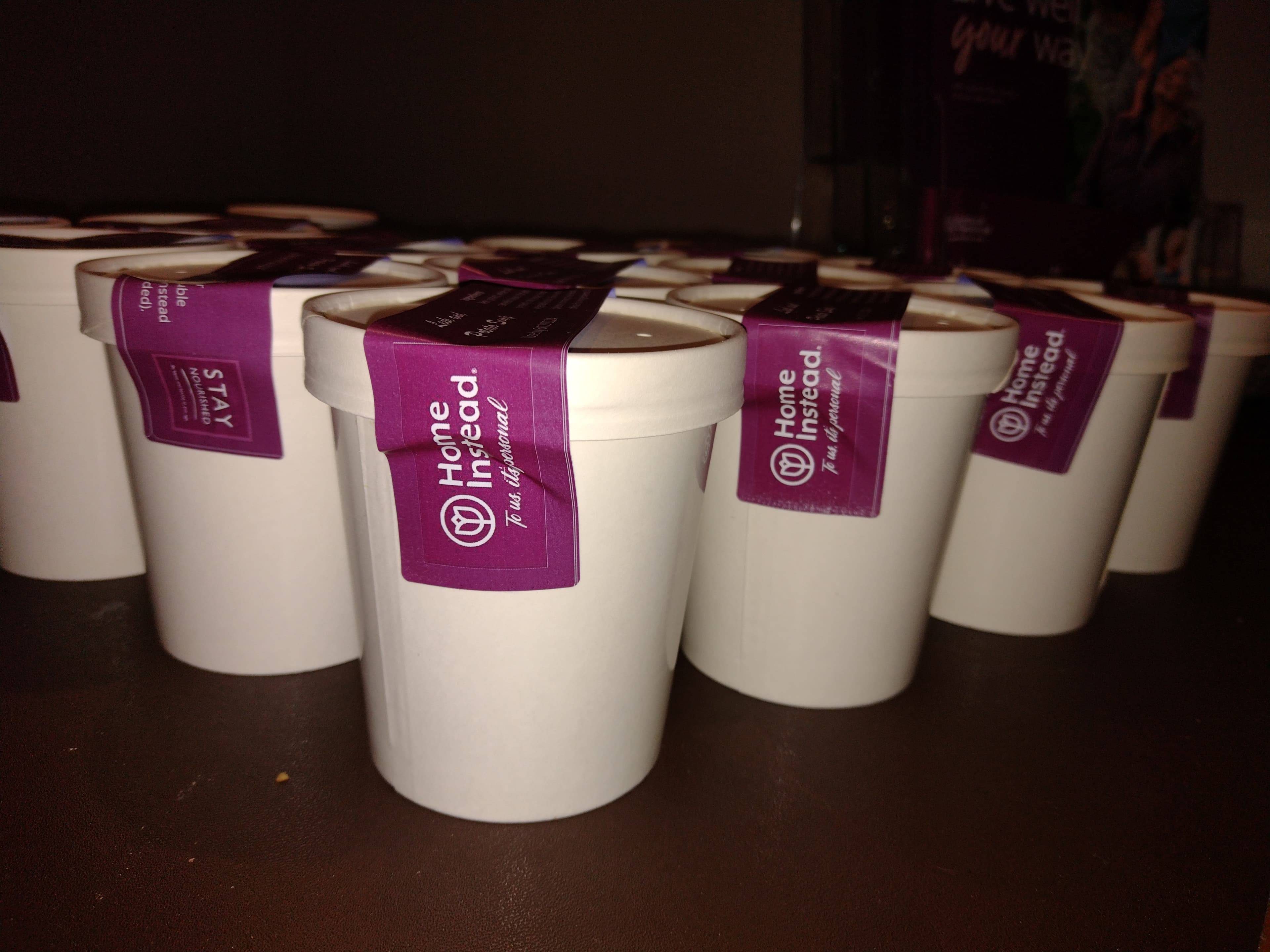 Multiple white paper cups with purple "Home Instead" stickers on their lids, arranged on a brown surface. - Home Instead