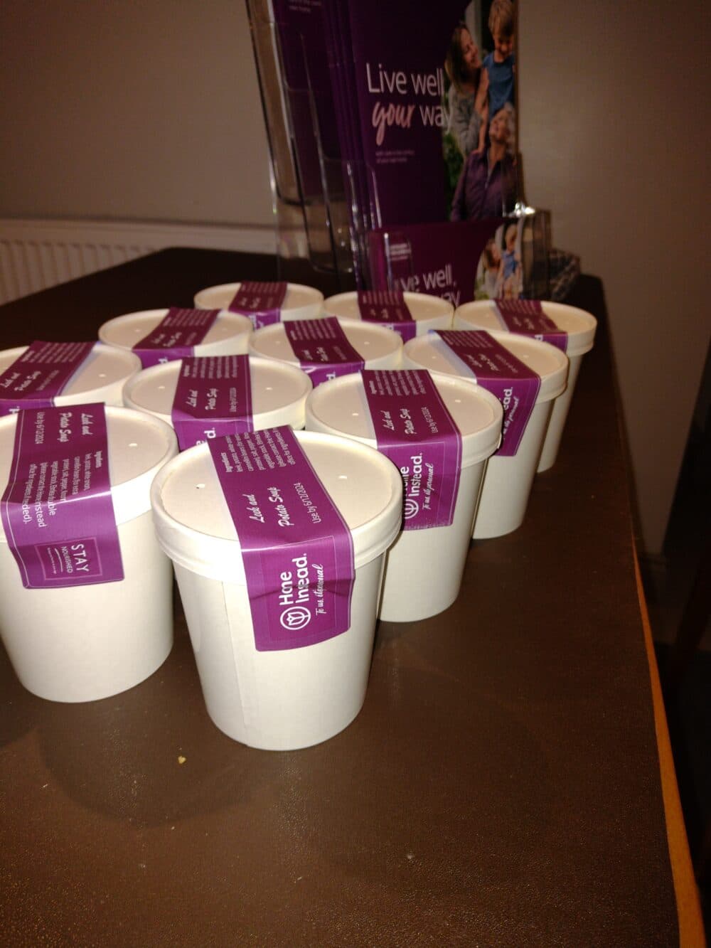 Several white paper cups with purple labels are arranged on a brown table near brochures. - Home Instead