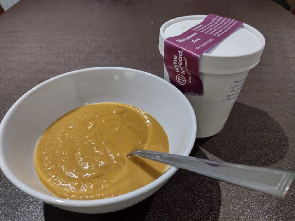 A bowl of thick orange soup with a spoon, next to a white takeout container on a brown table. - Home Instead