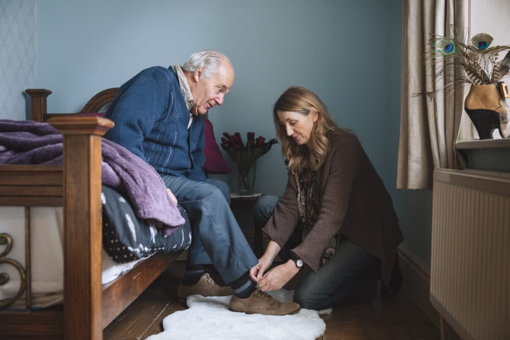A woman kneels to help an elderly man put on shoes beside a bed in a cozy, softly lit room. - Home Instead