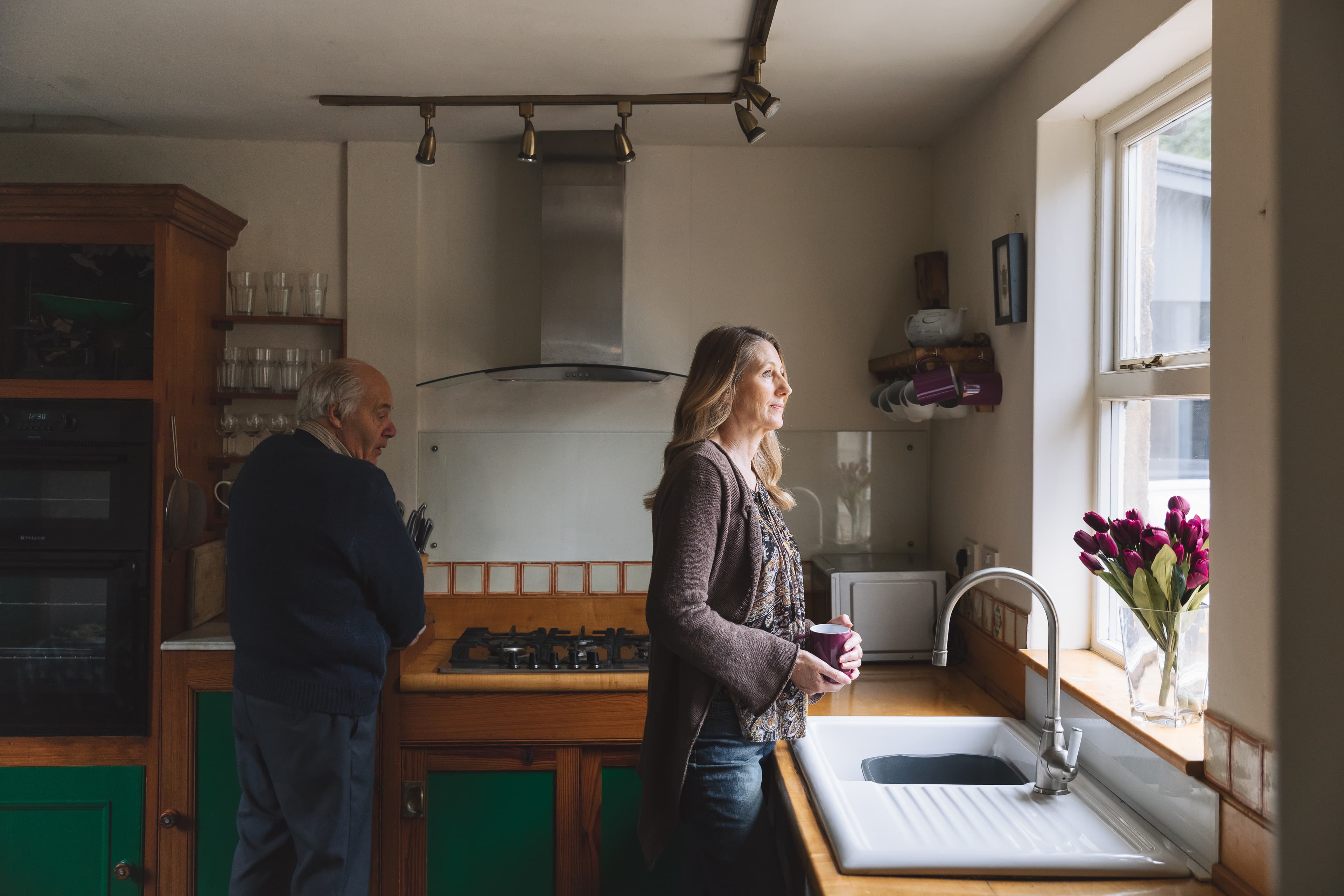 A woman with a mug looks out a kitchen window while an older man stands near the stove behind her. - Home Instead