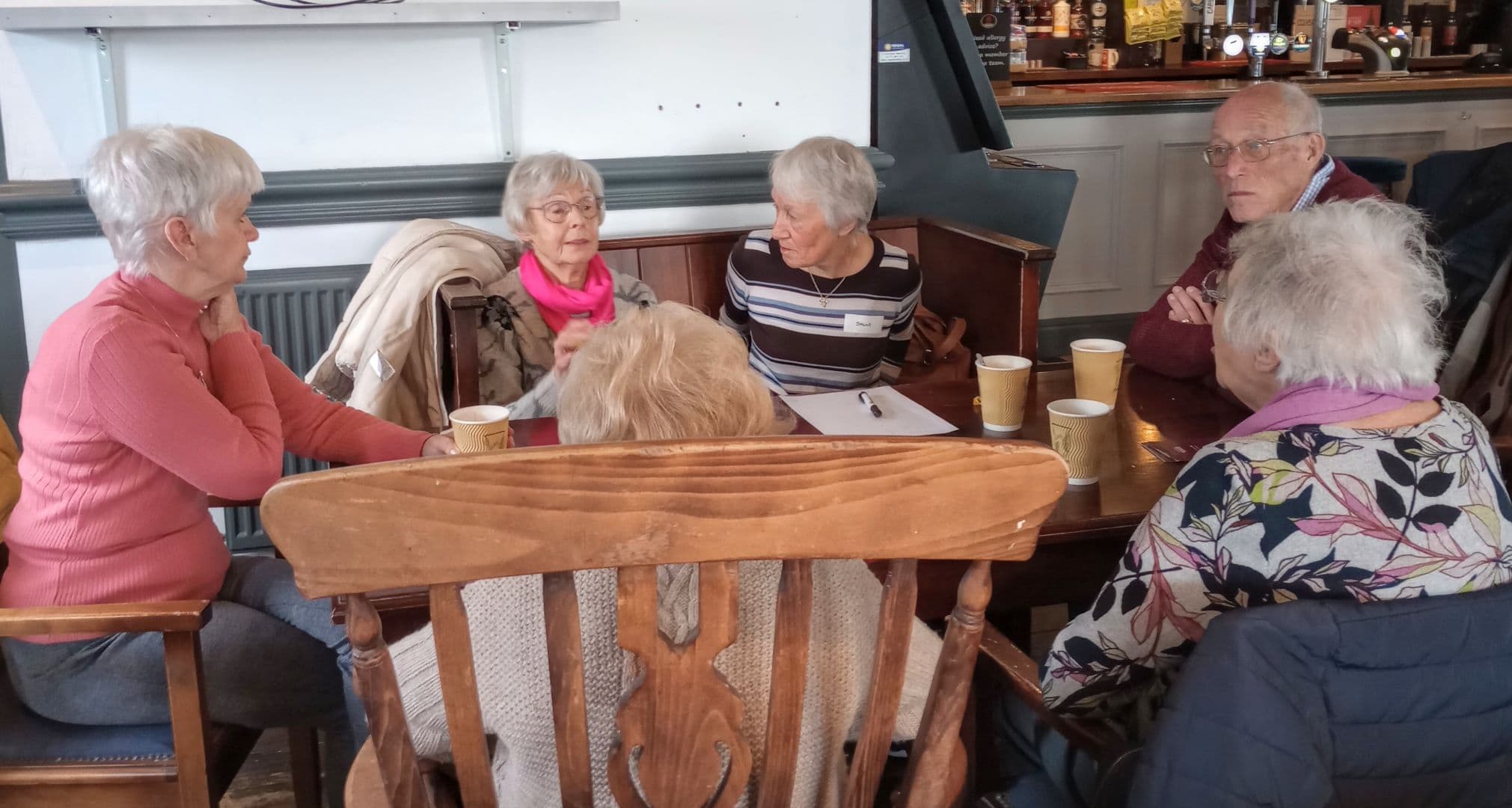 Six older adults sit around a wooden table in a café, talking with drinks in takeaway cups. - Home Instead