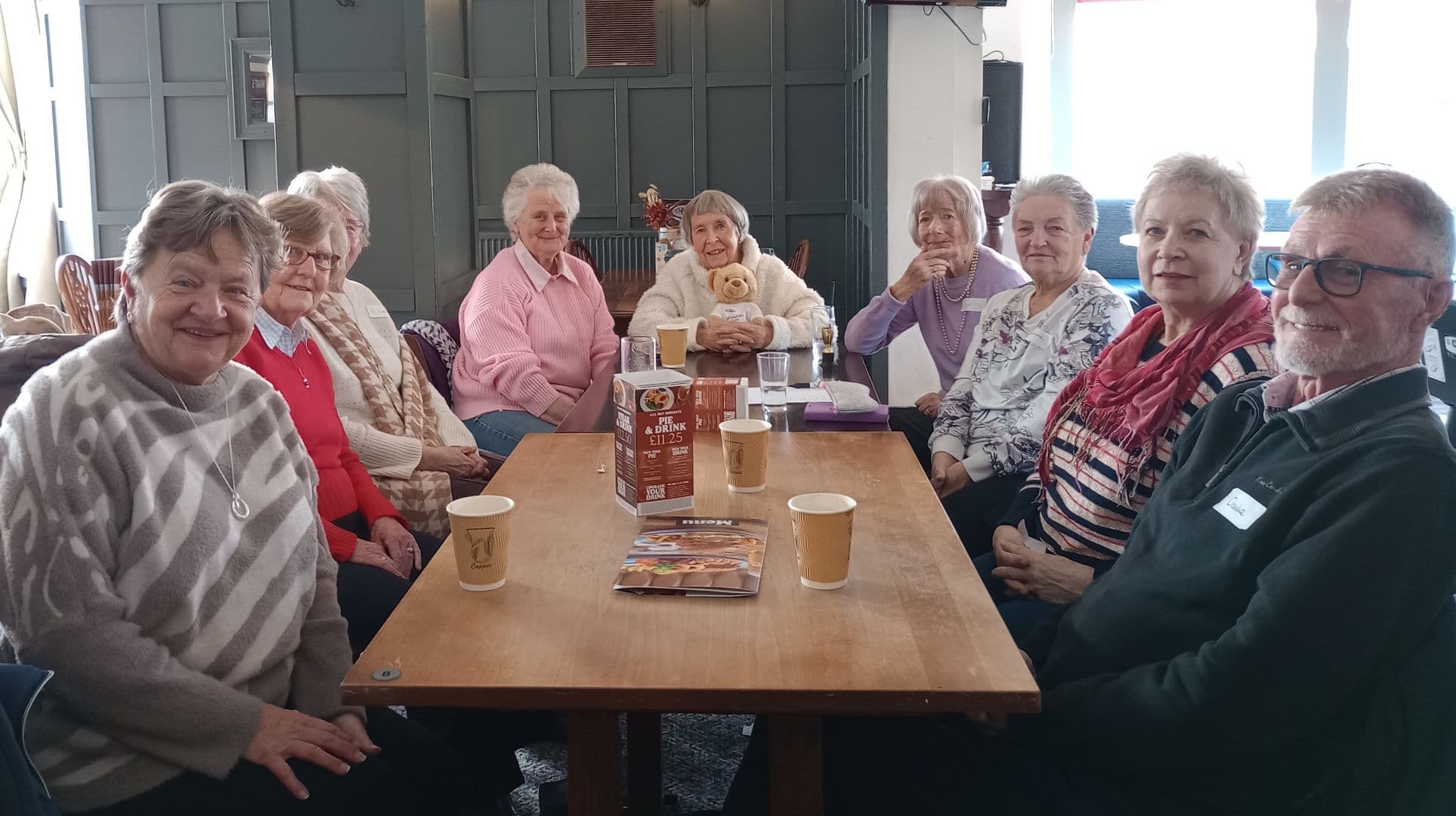 A group of older adults sit around a wooden table, smiling and enjoying drinks together in a cozy room. - Home Instead