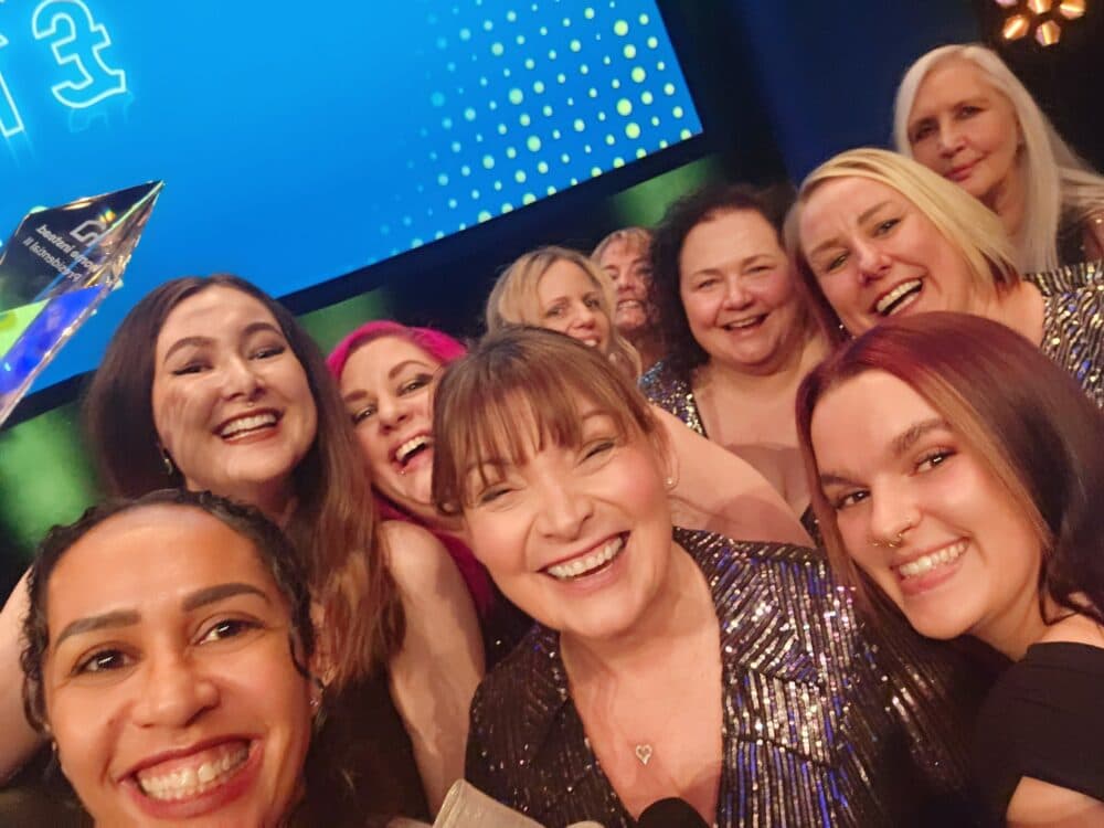 A group of smiling women in sparkly outfits take a selfie at an awards event, holding a trophy. - Home Instead
