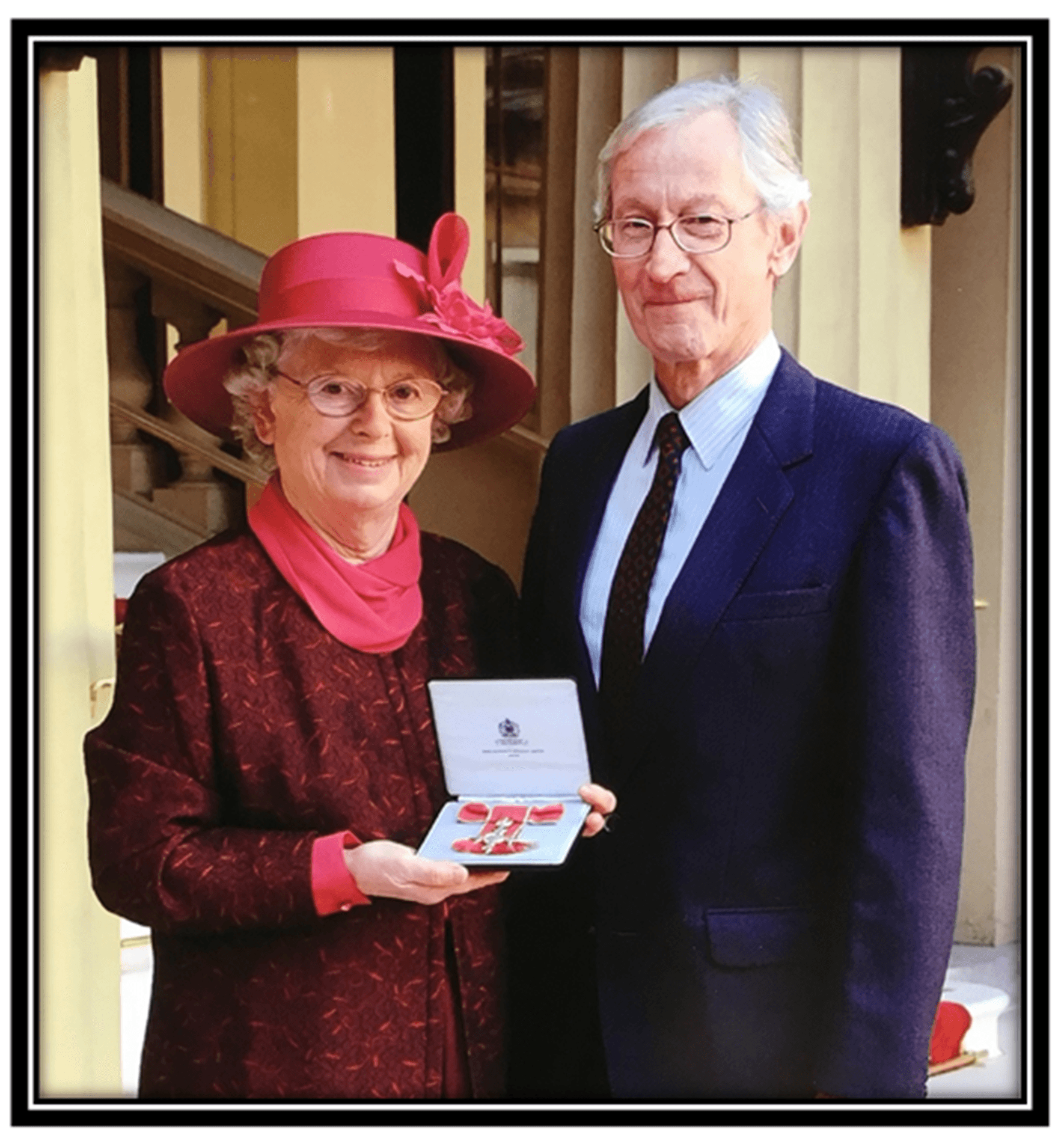 An elderly woman in a red hat holds an award beside an elderly man in a suit, both smiling. - Home Instead