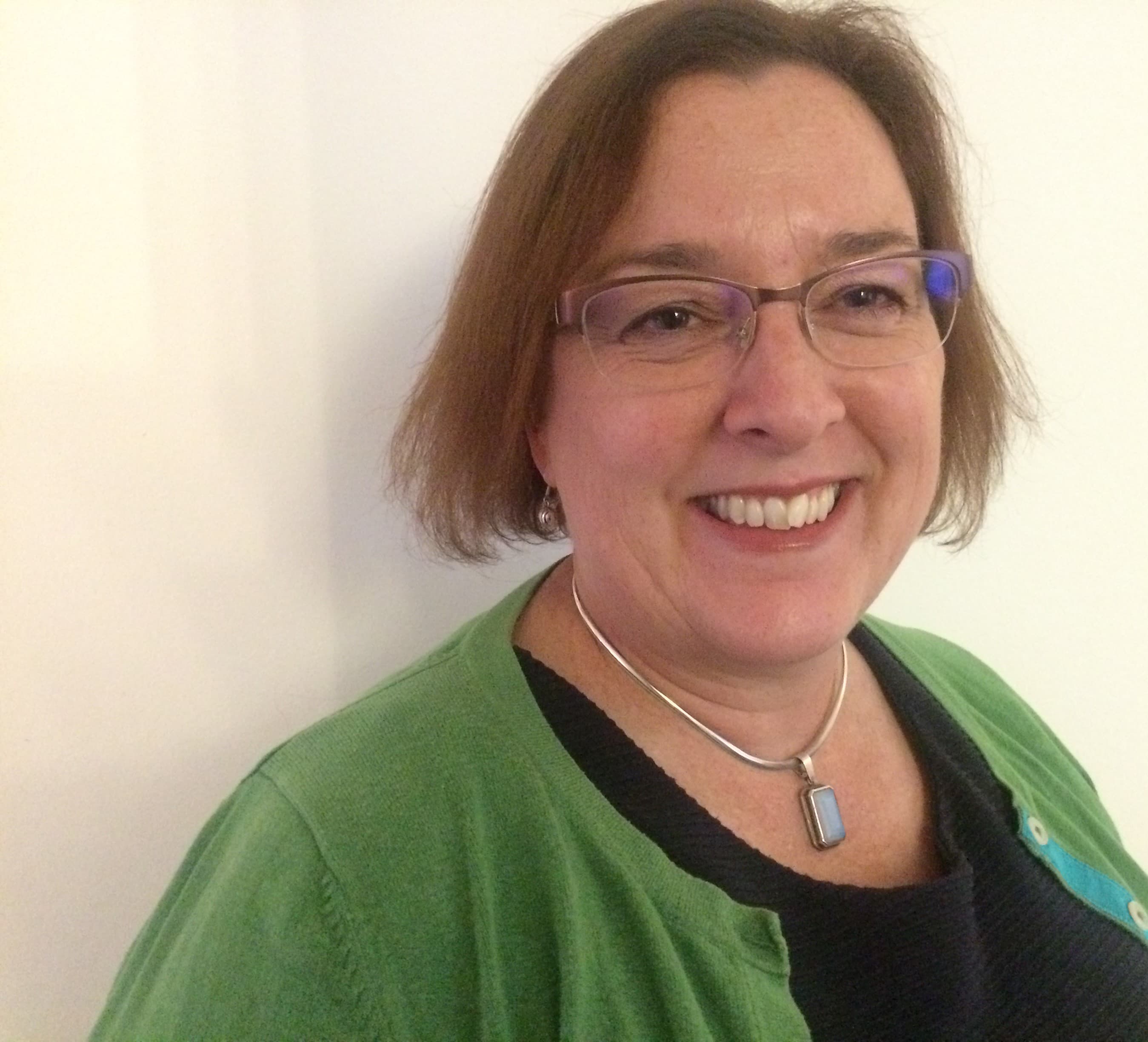 Smiling woman with glasses, short brown hair, and a green sweater stands against a plain light background. - Home Instead