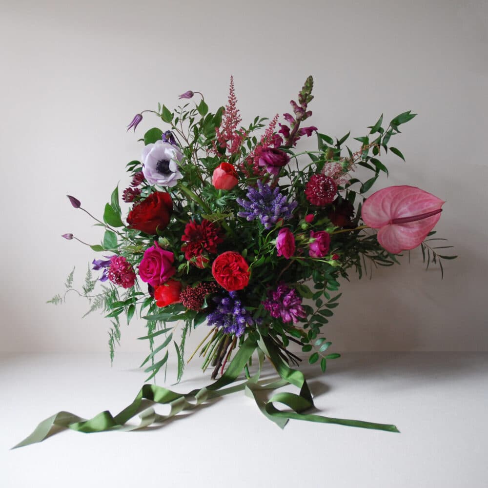 A lush, romantic bouquet featuring a mix of deep red, pink, and purple flowers, including roses, anemones, and snapdragons, accented by delicate greenery. A striking pink anthurium adds a bold touch, while long green ribbons flow from the base. The arrangement is set against a neutral white background.
