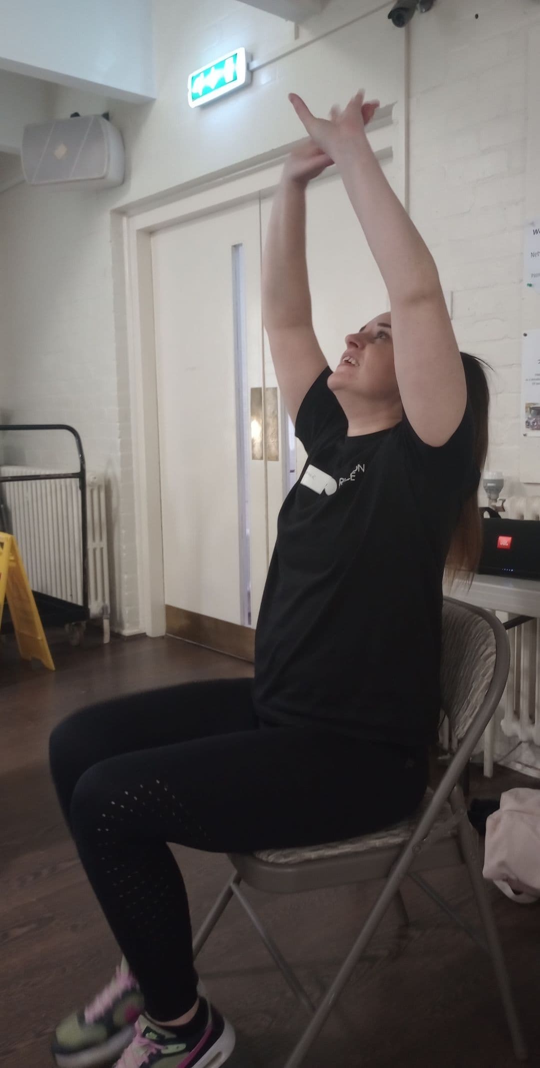 Woman sitting on a chair indoors, stretching both arms upward, wearing a black shirt and leggings. - Home Instead