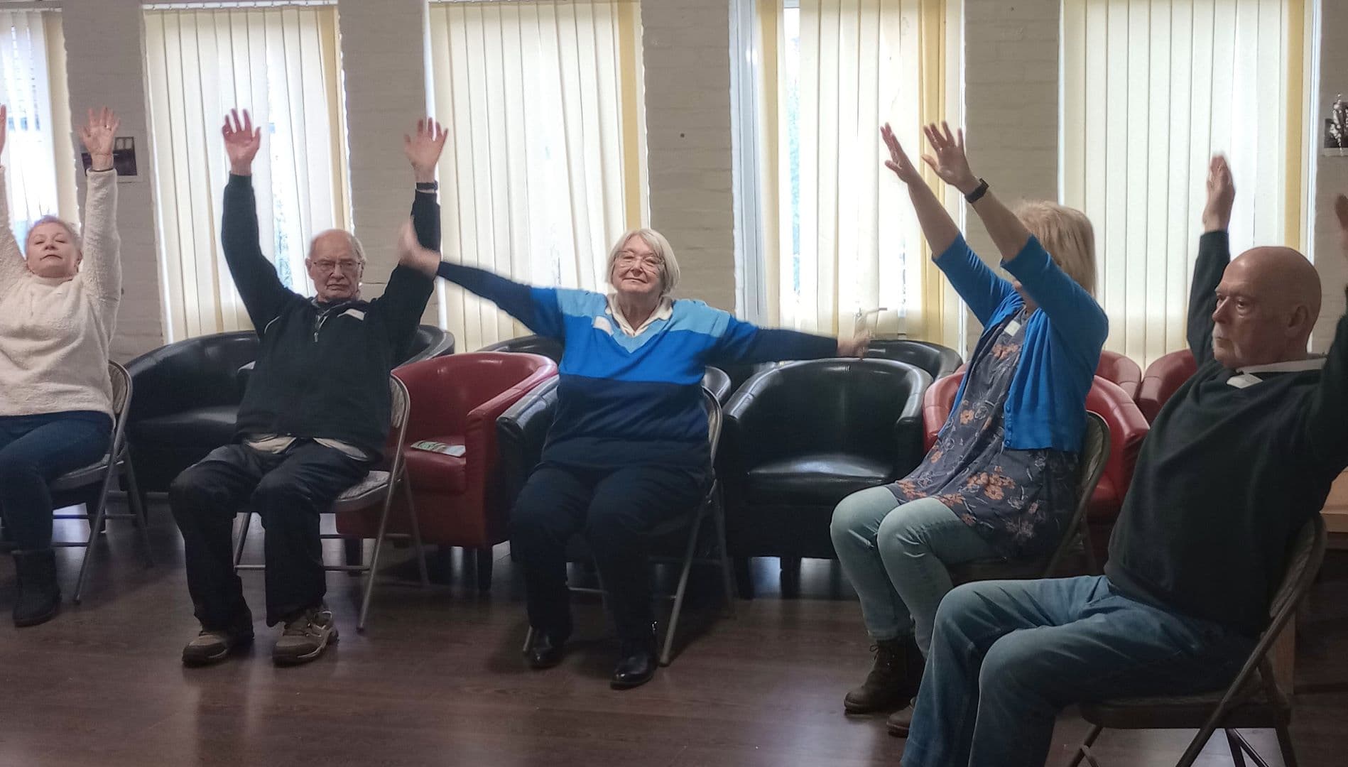 Five older adults sit on chairs in a row, raising their arms in a group exercise session. - Home Instead