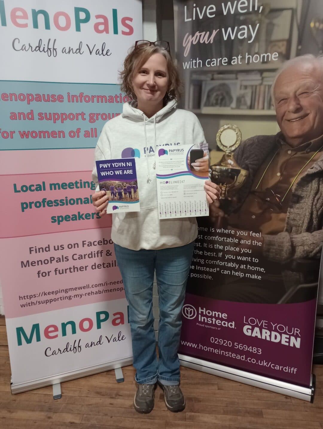 Woman in a hoodie holding leaflets, standing between two informative banners about support and care services. - Home Instead
