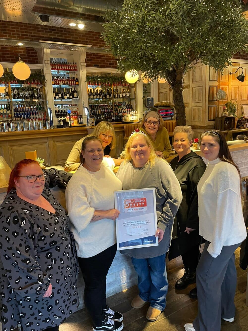 Six women pose together in a restaurant, one holding a framed certificate, with a bar and tree in the background. - Home Instead