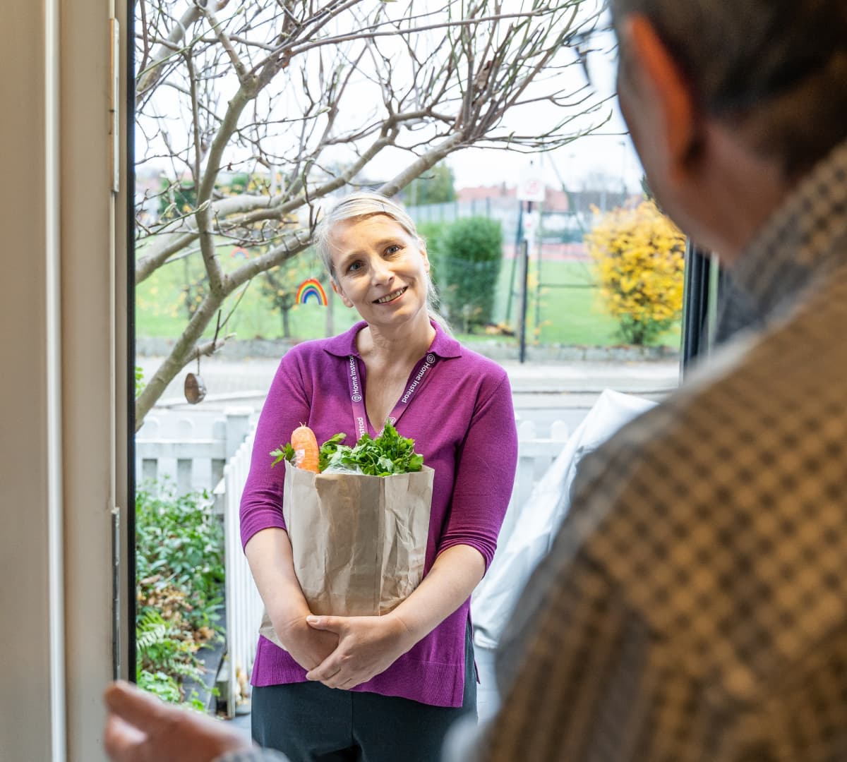 A female carer wearing purple and with her hair tied up at the door while bringing a bag of groceries