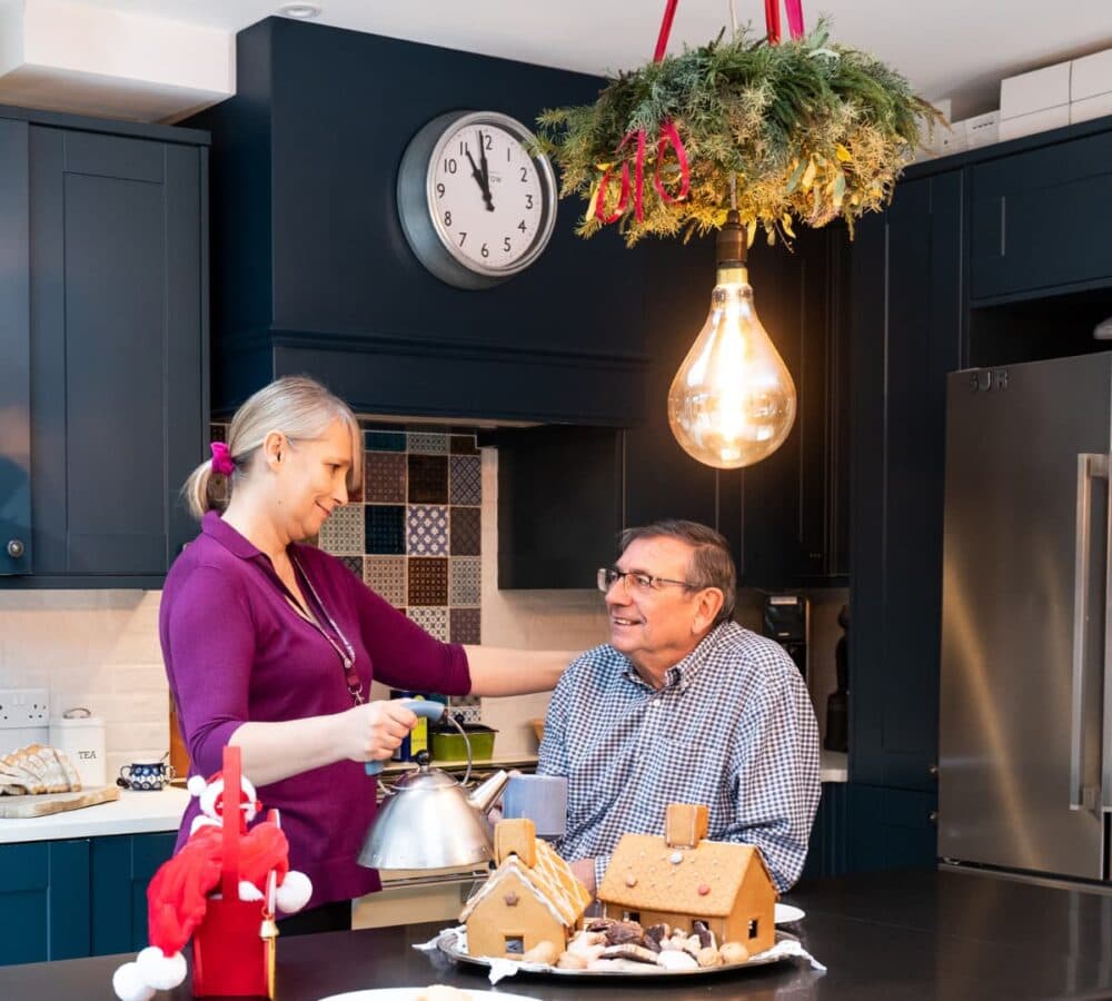 A female carer with her hair tied and wearing purple smilign while chatting with an male older adult with eyeglasses and wearing long sleeves inside the kitchen
