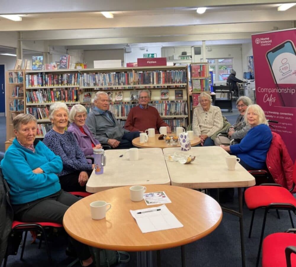 Group of older adults men and women all sitting on a chair with a table and all are smiling and happy inside the library
