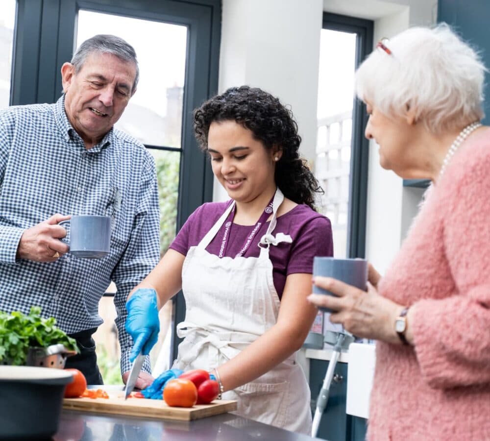 Two older adults with grey hair inside the kitchen while drinking coffee with a younger female carer with long black curly hair chopping vegetables