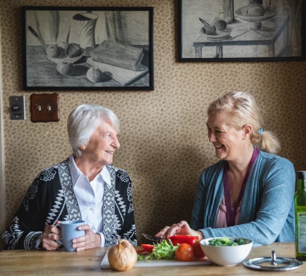 Younger female carer cutting vegetables while chatting with a female older adult with grey hair inside the kitchen