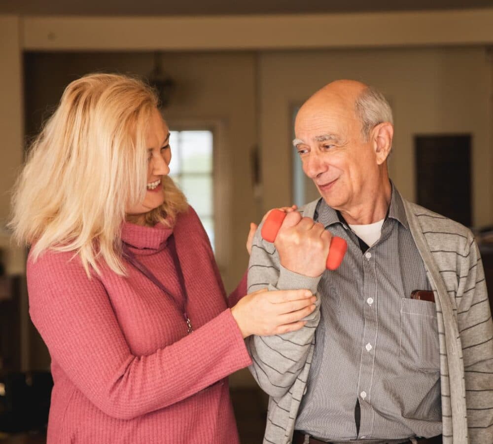 Older man with grey hair exercising at home with his carer with long blonde hair and wearing pink assisting him