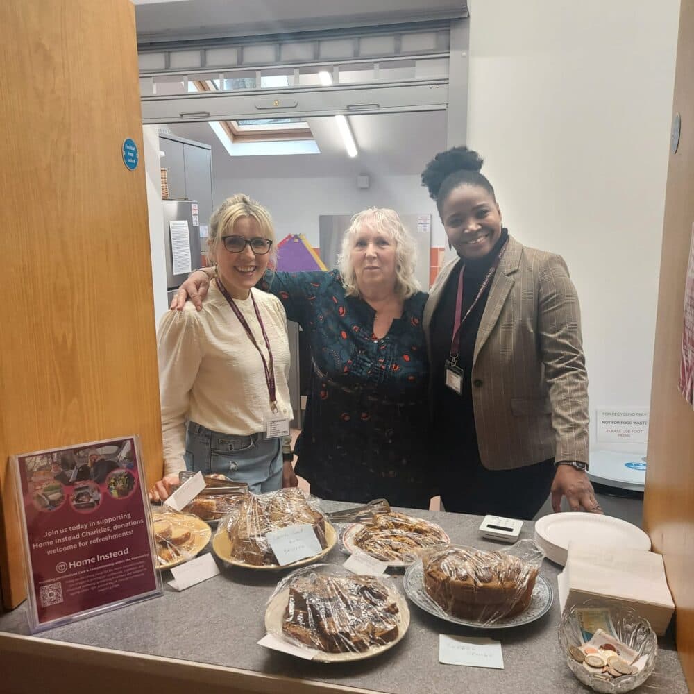 Three women stand behind a counter with assorted cakes and baked goods on display at an indoor event. - Home Instead